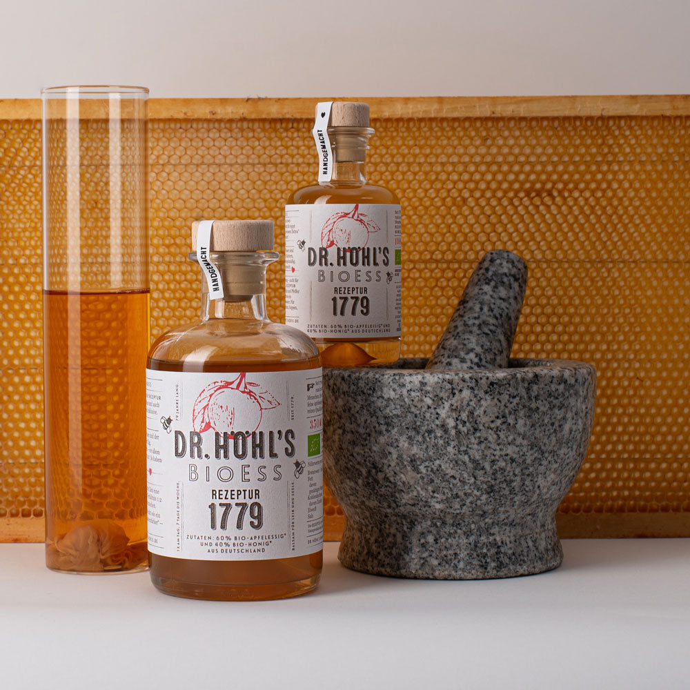 Reagenzglas mit rohem Apfelessig zwei Flaschen REZEPTUR 1779 und ein Mörser vor einer Honigwabe