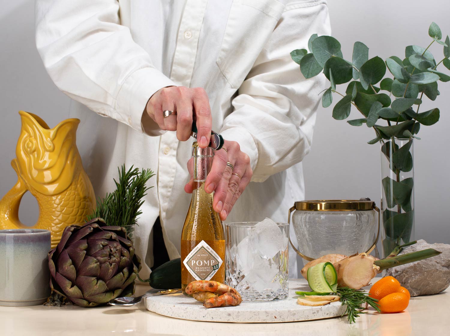 frau öffnet eine piccolo flasche alkoholfreier aperitif feinherb, die neben einem glas mit eiswürfeln und dekoration steht