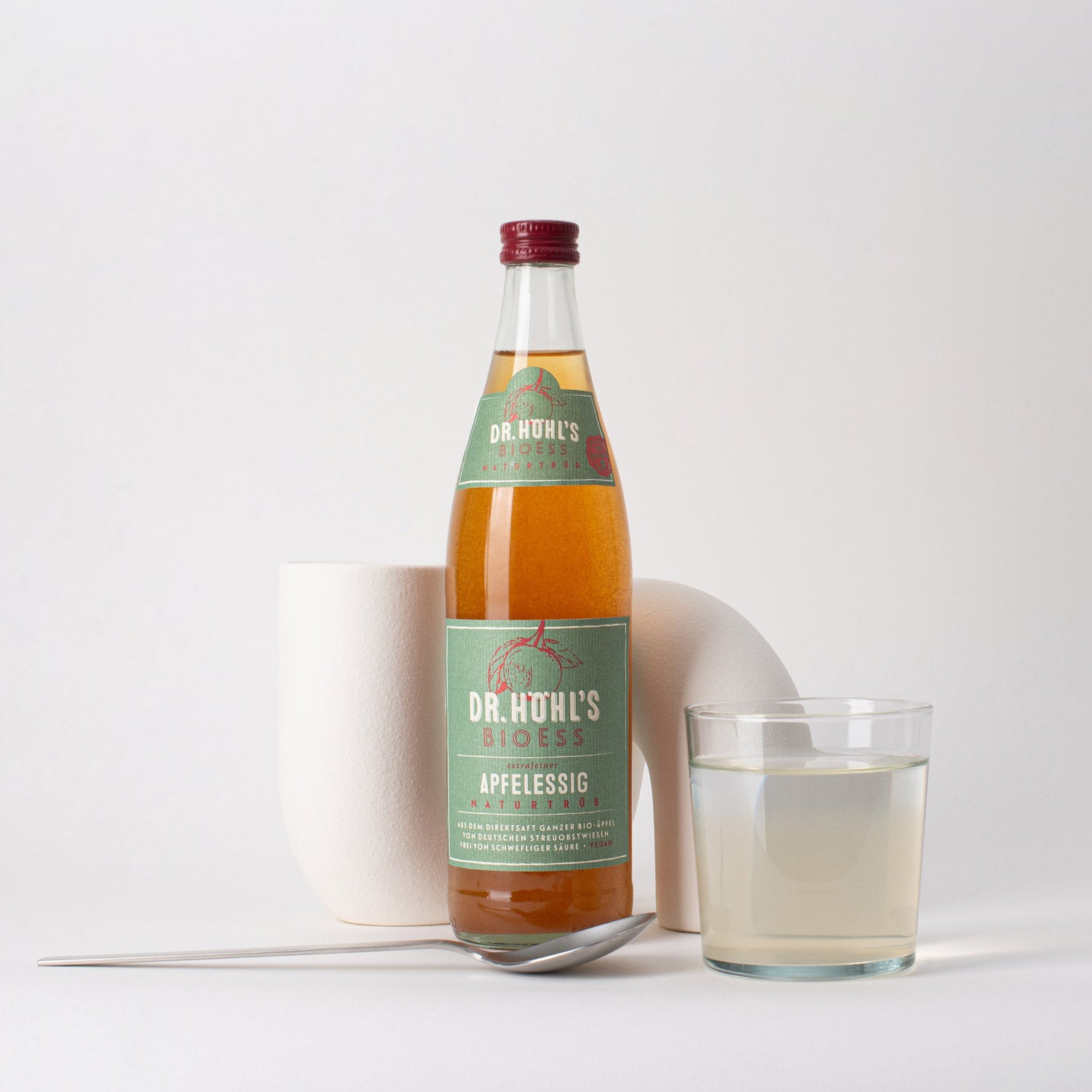 helles stillleben einer flasche bio-veganer naturtrüber bioess apfelessig mit einem esslöffel sowie einem glas wasser mit apfelessig