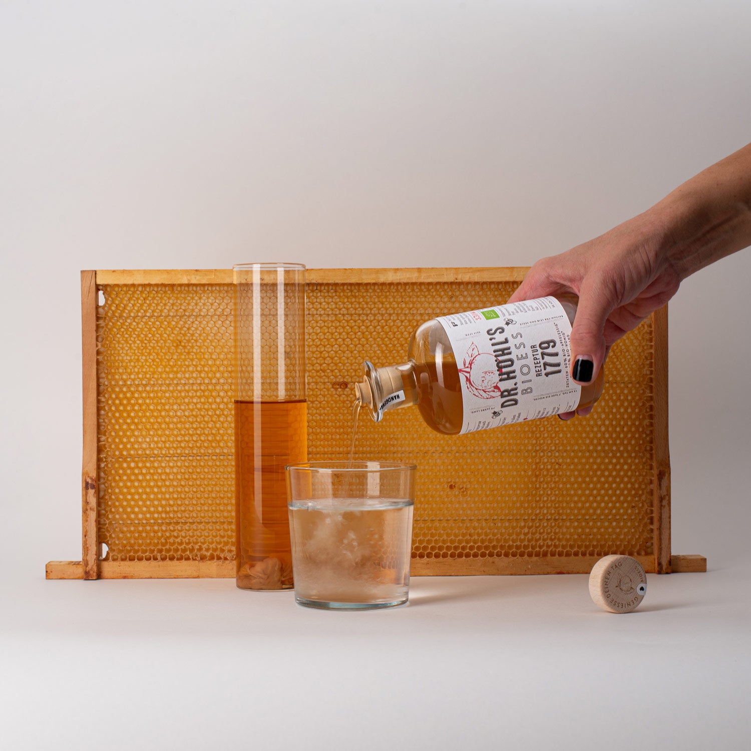 stillleben mit echtem honig waben rahmen, davor eine apothekerflasche rezeptur 1779 von einer hand gehalten beim einschenken der apfelessig honig mischung in ein glas wasser , sowie ein glaszylinder mit apfelessig mutter