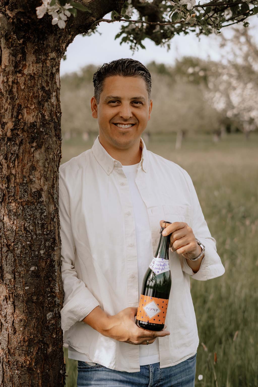 johannes lehnt an einen apfelbaum auf der streupbstwiese und hält eine flasche pomp klassik sekt in den händen