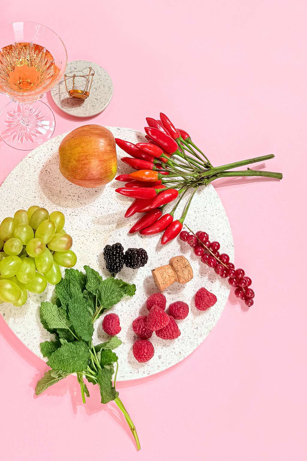 natuerliche zutaten wie apfel traube chili melisse und johannisbeere auf einem steintablett neben einem glas pomp rose alkoholfreier aperitif