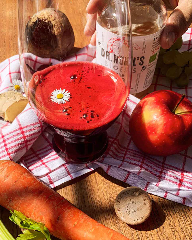 Roter Beete Karotten Sellerie Smoothie in einem Glas mit Gänseblümchen dekoriert und einer Flasche Apfelessig-Honig Mixtur